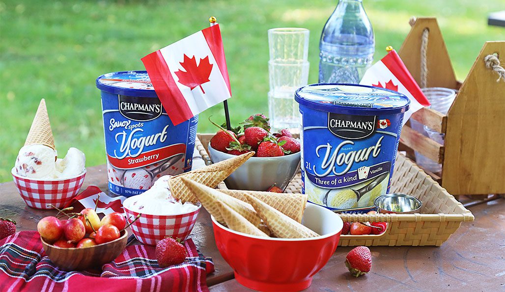 Outside with green grass in the background is a Canada Day themed spread on a wooden table. On the table are two tubs of Chapman's Frozen Yogurt, red bowls with fresh strawberries and waffle cones, and red and white checkered bowls with scoops of frozen yogurt.
