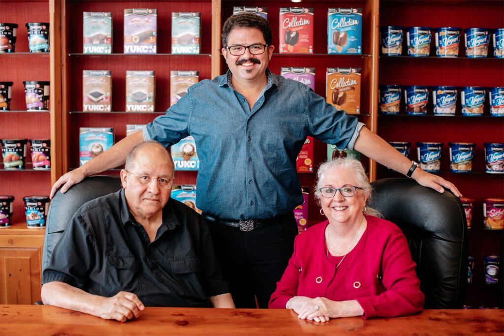 With Ashley Chapman standing behind them, David and Penny Chapman are sitting at a table in Chapman's head office boardroom. An array of Chapman's products are on display behind them.